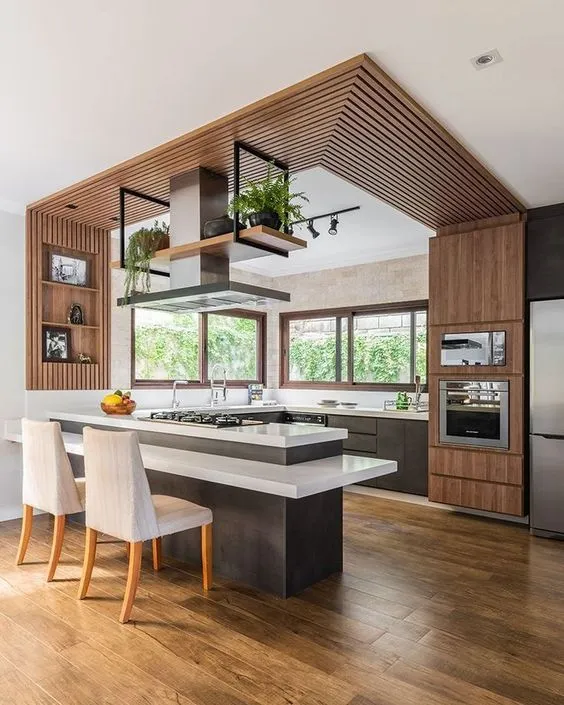 Modern kitchen with wood ceiling panel, dark island, two white chairs, oven, and wood floor