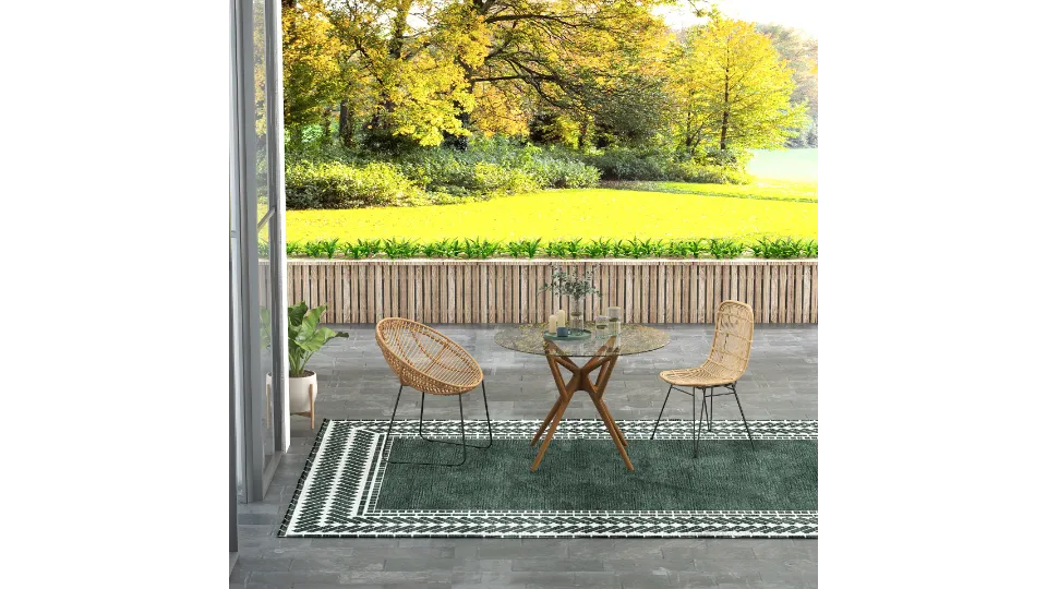 Outdoor patio featuring a glass table, two woven chairs, green rug, and sunny green garden