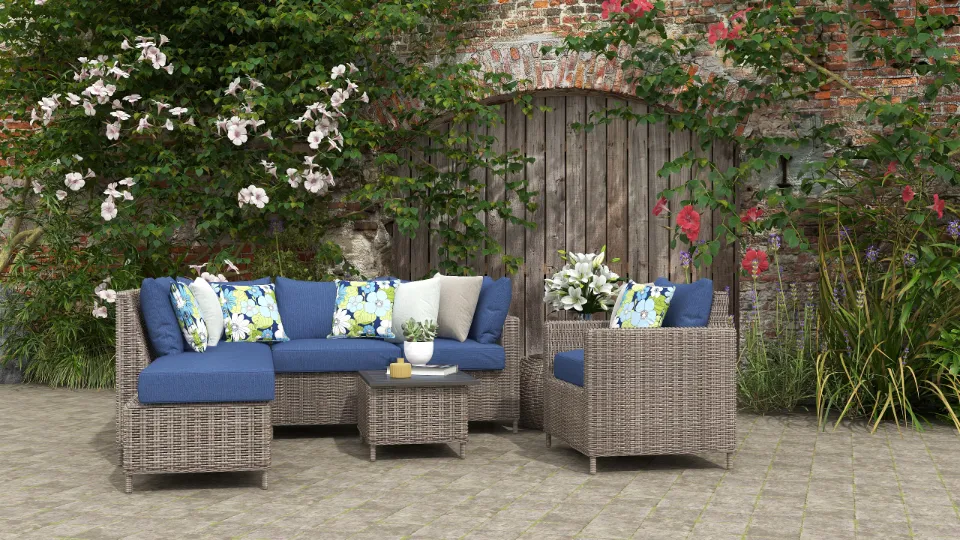 Outdoor patio with grey wicker sofa set, blue cushions, floral pillows, and flowering garden wall