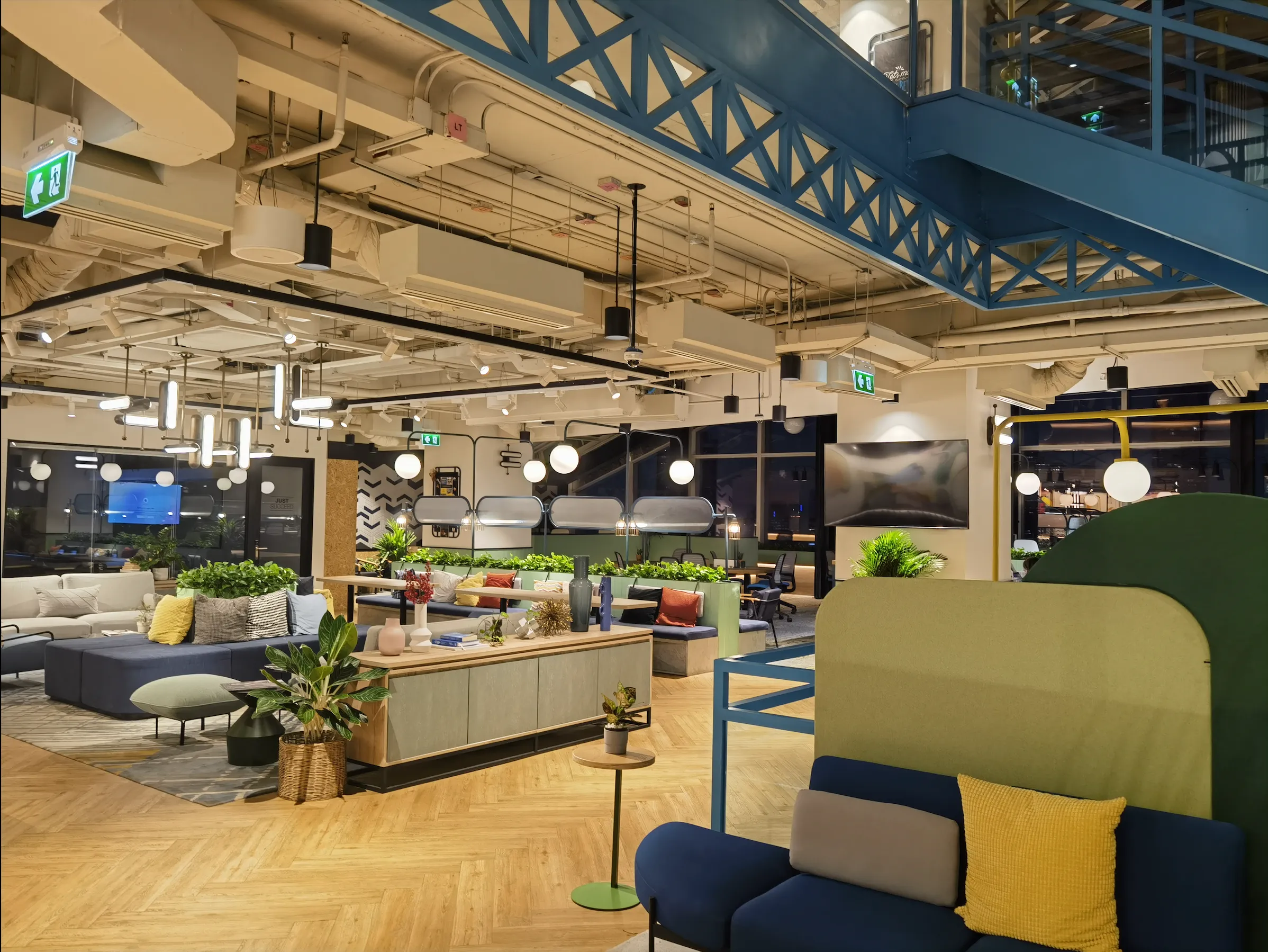 Modern office interior with blue sofas, colorful cushions, industrial ceiling, wooden floor, and green plants
